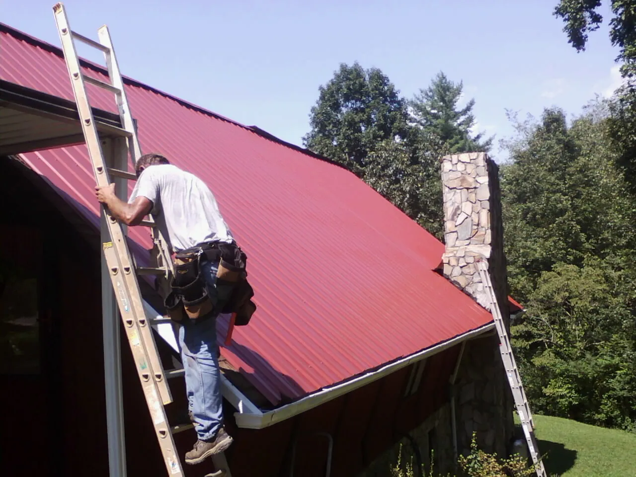 Roofer on a ladder beside a red metal roof and stone chimney — Soffit & Fascia Repair by Roofing Brothers in Barney